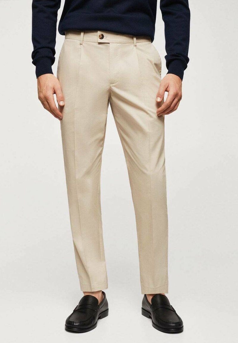 Pantaloni beige con vestibilità sartoriale, dotati di design a gamba dritta senza pieghe, tasche frontali e chiusura con un solo bottone. Indossati con mocassini neri.