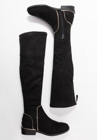 Sorte over-knee støvler lavet af ruskind, udstyret med en guldaccent lynlås og syningsdetaljer. Flad sål med en let hæl.