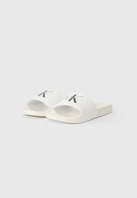 Λευκά σανδάλια με πλατιά υφασμάτινη λωρίδα που διαθέτει μαύρο λογότυπο. Κάτω μέρος σε επίπεδο, ορθογώνιο σχήμα με λεία υφή, μινιμαλιστικός σχεδιασμός.