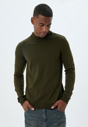 Olivengrüner Rollkragenpullover aus leichtem Strick. Figurbetontes Design mit langen Ärmeln und gerippten Bündchen. Getragen mit Jeans.