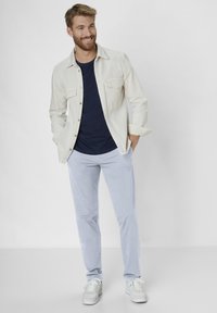 Men's outfit featuring a cream jacket, navy t-shirt, light blue pants, and white sneakers with textured soles. Jacket has buttoned pockets.
