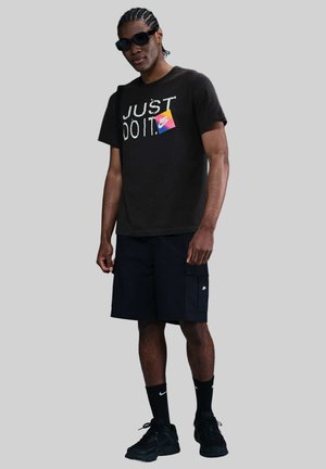 Maglietta nera con grafica "JUST DO IT", pantaloni cargo neri e sneakers nere. Gli accessori includono occhiali da sole e calzini neri.