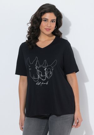 Femme aux cheveux bouclés et foncés portant un T-shirt noir à col en V affichant un dessin au trait blanc de trois visages d'animaux et le texte "meilleurs amis".