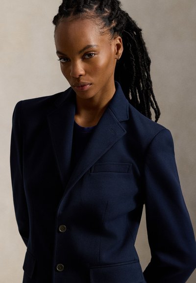 Γυναίκα με μακριά πλεξούδα που φοράει ένα σκουρομπλε σακάκι πάνω από ένα σκουρόχρωμο τοπ σε απλό ουδέτερο φόντο.