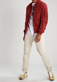 Czerwona kurtka bombowca o gładkiej teksturze, z zamkiem błyskawicznym i kieszenią na rękawie; połączona ze kremowymi spodniami i beżowymi sneakerami z białymi akcentami.