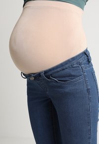 Fascia di supporto per la pancia in maternità beige indossata sotto una maglietta aderente, con jeans blu scuro parzialmente slacciati in vita, caratterizzati da un bottone e dettagli delle tasche.