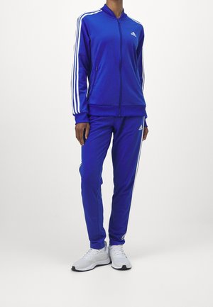 Persona che indossa una tuta Adidas blu con strisce bianche sulle maniche e sui pantaloni, abbinata a scarpe da ginnastica bianche, in piedi davanti a uno sfondo bianco.