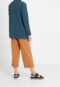 Blazer azul petróleo de manga larga, combinado con pantalones cortos naranjas y sandalias negras con suelas blancas. 100% algodón, corte relajado, diseño minimalista.