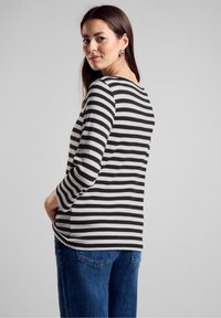 Pasek bluzka z długim rękawem w czarno-białe paski, wykonana z miękkiego materiału, okrągły dekolt, boczne rozcięcia, zestawiona z niebieskimi dżinsami z tylnymi kieszeniami.
