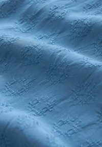 Texturovaná modrá látka s vyvýšenými geometrickými vzory a hladkým povrchem. Materiál má jemný, lehký vzhled.