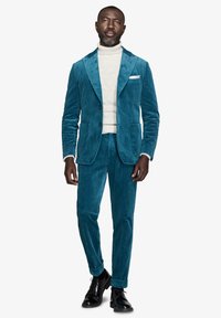 Teal corduroy-puku, jossa on lovettu kaulus, yhdistettynä vaalean cremeen poolopaitaan. Mustat kengät viimeistelevät asun. Sujuva tekstuuri, tarkka istuvuus.