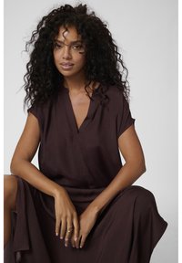 Robe à manches courtes, de couleur bourgogne profond, avec un décolleté en V, en tissu doux et texturé, et une silhouette fluide. Le modèle pose assis, les mains reposant sur les genoux.