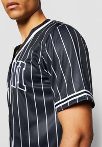 Čierna baseballová dres so bielymi vertikálnymi pruhmi, v-krkom a krátkymi rukávmi s kontrastným lemovaním. Na prednej strane veľké písmo.