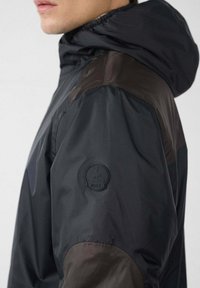 Man draagt een zwart-bruine jas met capuchon, met een geborduurd rond logo op de bovenkant van de mouw.