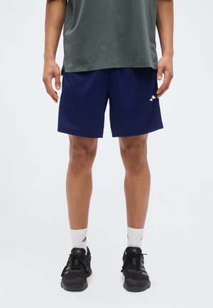Bărbat purtând pantaloni scurți sportivi bleumarin, un tricou gri, șosete albe și pantofi de alergare negri Adidas, stând în fața unui fundal simplu.