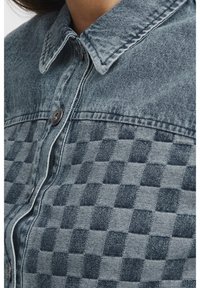 Koszula dżinsowa z kołnierzykiem zapinanym na guziki. Charakteryzuje się teksturowanym wzorem w kratę w różnych odcieniach niebieskiego i szarości oraz srebrnymi guzikami.