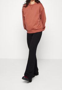 Persona vestida con un suéter color óxido, pantalones acampanados negros con texturizado y zapatos negros gruesos con detalles burdeos, de pie contra un fondo blanco.