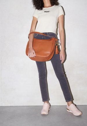 Bolso de cuero color canela con textura suave y detalle de borla, combinado con vaqueros oscuros y zapatillas de color rosa claro, sobre un fondo neutro.