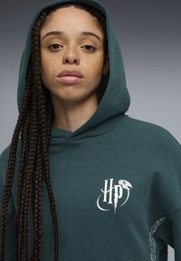 Sudadera con capucha de tejido verde oscuro con logo gráfico blanco en el pecho. Presenta una textura suave y acentos de trenzas de cabello.