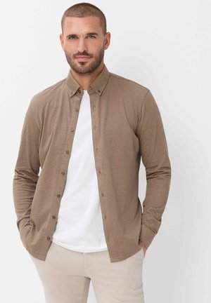 Mand med kort hår og skæg iført en lysebrun skjorte med knapper over en hvid T-shirt og beige bukser, stående med hænderne i lommerne.