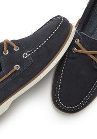 Marineblaue Wildlederschuhe mit weißer Naht, braunen Schnürsenkeln und Gummisohle. Sie verfügen über ein klassisches Bootsschuh-Design mit Metallösen.