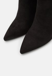 Zapatos de tacón negros de ante con punta afilada y una textura suave. El diseño presenta una silueta elegante adecuada tanto para ocasiones formales como informales.
