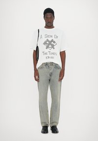 White cotton t-shirt with graphic print, paired with light blue denim jeans. Black shoes and a small black shoulder bag. Neutral background.