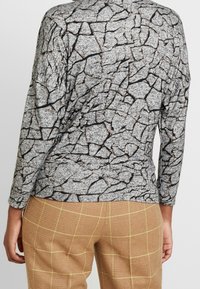 Camiseta de manga larga en tejido gris con un patrón de grietas texturizado en negro y acentos marrones. Combinada con pantalones de cuadros marrones.