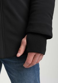 Juodas švarkas su tekstūruotu rankogaliu. Lygus audinys su prigludusia siluetu, derinamas su mėlynomis džinsinėmis kelnėmis. Rankos poilsis ant klubų.