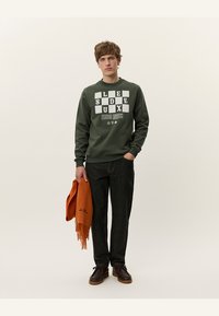 Sudadera verde con texto y gráficos blancos, jeans oscuros, zapatos marrones, sosteniendo una bufanda naranja con flecos. Atuendo simple y casual.