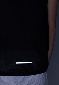 Černé sportovní tričko s texturovaným, prodyšným designem. Má reflexní vodorovný pruh blízko lemu, který kontrastuje s hladkým materiálem.