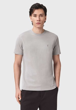Ung mand iført en ensfarvet lysegrå kortærmet rundhalset t-shirt med et lille logo på venstre bryst, stående med den ene hånd i lommen.