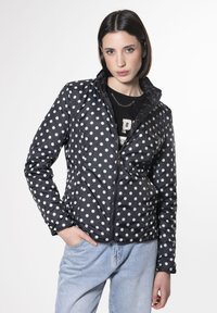 Chaqueta negra acolchada con grandes lunares blancos, cierre de cremallera, cuello alto y bolsillos laterales, combinada con jeans de color azul claro.