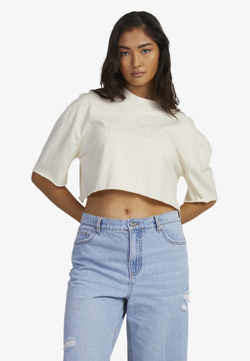 Cropped cream cotton t-shirt with a ribbed crew neck and short sleeves; features a subtle, embossed logo in the center. Paired with light blue jeans.