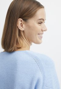 Svetlo modri pleteni pulover z izdelano teksturo, širokim ovratnikom in vidnimi šivi, prikazan iz stranske perspektive.