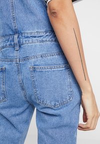 Salopette en denim bleu clair. Présente une taille cintrée, des jambes droites, des poches arrière et un tatouage sur le bras tenant la bretelle.