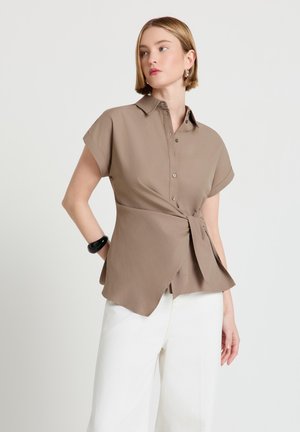 Woman with short blonde hair wearing a brown short-sleeve buttoned blouse tied at the waist and white pants, standing with one hand on hip.