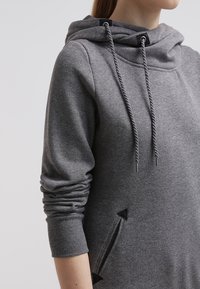 Harmaa huppari pehmeästä kankaasta, jossa on kaulus, kiristysnyöri ja sivutaskut, joissa kolmiomaiset leikkauskoristeet. Pitkät, istuvat hihat.