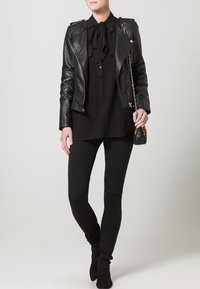 Zwarte leren motorjack met decoratieve ritsen, over een zwarte blouse met een strik-kraag. Gecombineerd met zwarte slim-fit broeken en enkelboots.