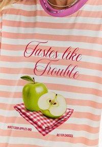 Pruhované tričko v ružovej a bielej farbe. Obsahuje grafiku zeleného jablka a rozpoleného jablka na červenom kockovanom ubruse, so dekoratívnym textom.