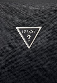 Black textured material featuring a triangular metal logo with “GUESS USA” text and a question mark design.