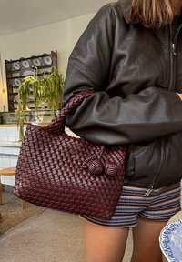 Borsa in tessuto color bordeaux a forma arrotondata, con due accenti a forma di pallina testurizzati e manici solidi, abbinata a una giacca di pelle scura e pantaloni corti a righe.