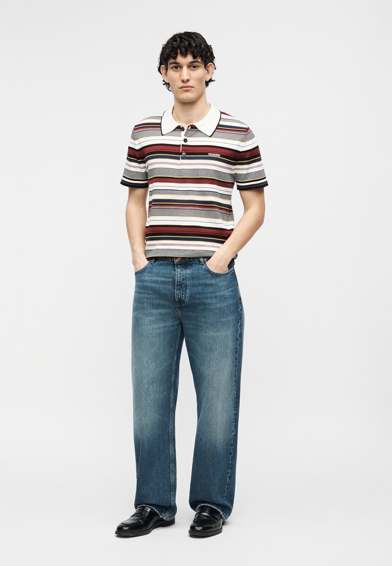Young man standing with hands in pockets wearing a striped short-sleeve polo shirt, blue jeans, and black shoes against a plain background.