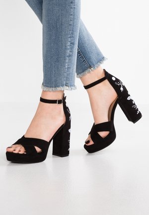 Füße, die schwarze Sandalen mit hohen Absätzen, Knöchelriemen und weißer Blumenstickerei tragen, kombiniert mit blauen Jeans mit ausgefranstem Saum.