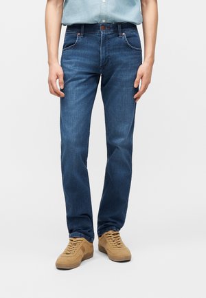 Personne portant un jean bleu à jambes droites, une chemise bleu clair rentrée, et des baskets beiges à lacets, debout devant un fond uni.