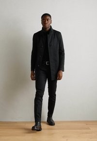 Zwarte jas met staande kraag, twee knopen en een matte textuur, gedragen over een zwart shirt en broek, gecombineerd met glanzende zwarte schoenen.