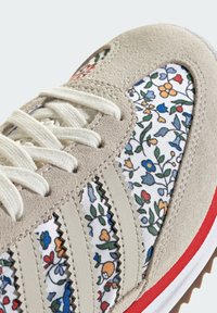 Sneakers med en beige mockaovandel, sidopanel i blommönstrad tyg, vita skosnören och röd gummi-sula. Har randig dekoration.