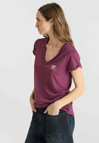 Vrouw met lang haar, gekleed in een kortmouwige bordeauxrode V-hals shirt en donkere denim broek, staande met handen in de zakken, kijkend naar links.