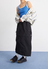 Blå ribbad tanktop med vit kant, oversized beige jacka med blå linjer, svart cargkjol och svarta sneakers med gula accenter.