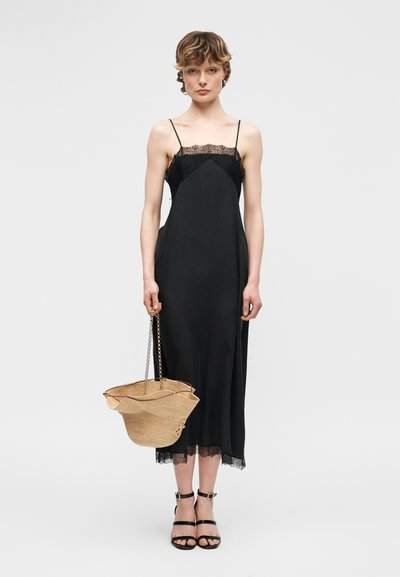 Γυναίκα με μαύρο φόρεμα αμάνικο με δαντέλα, φοράει μαύρα πέδιλα με λουράκι στον αστράγαλο, κρατώντας μια μεγάλη μπεζ υφαντή τσάντα με αλυσίδες ως χερούλια.